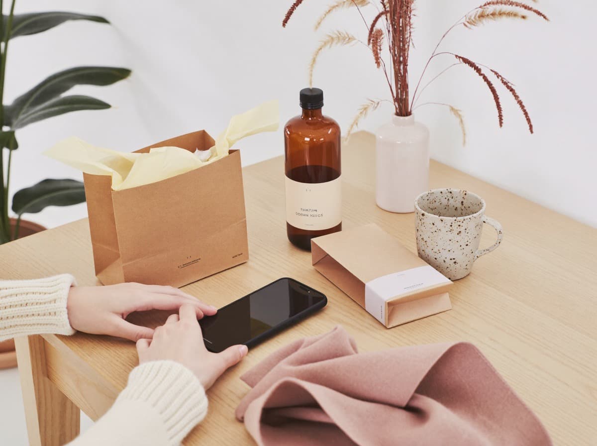Uma criadora abre um pacote de marca na mesa, com o celular na mão, ao lado de um frasco e uma sacola kraft